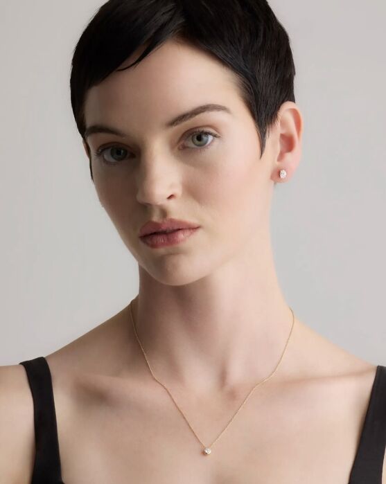 Woman with short dark hair wearing a black top, a small pendant necklace, and stud earrings, looking at the camera against a neutral background.