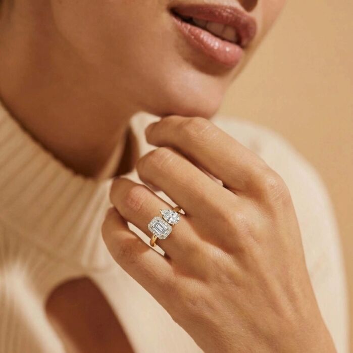 A person wearing a beige sweater displays a hand with a gold ring featuring three large clear gemstones, including an emerald cut center stone.