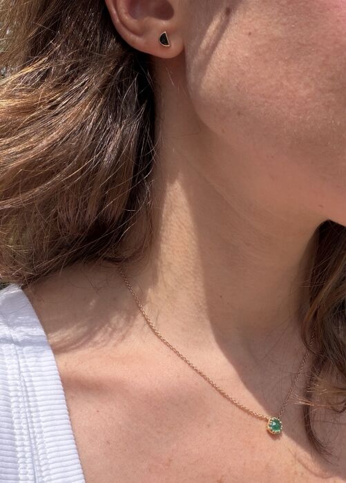 A woman wearing a small black triangular stud earring and a gold necklace with a green pendant, shown from the neck up.