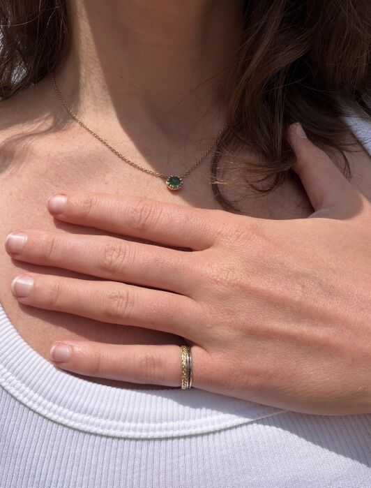 A person in a white tank top rests their hand on their chest, showing a gold and silver ring and a necklace with a green gemstone pendant.