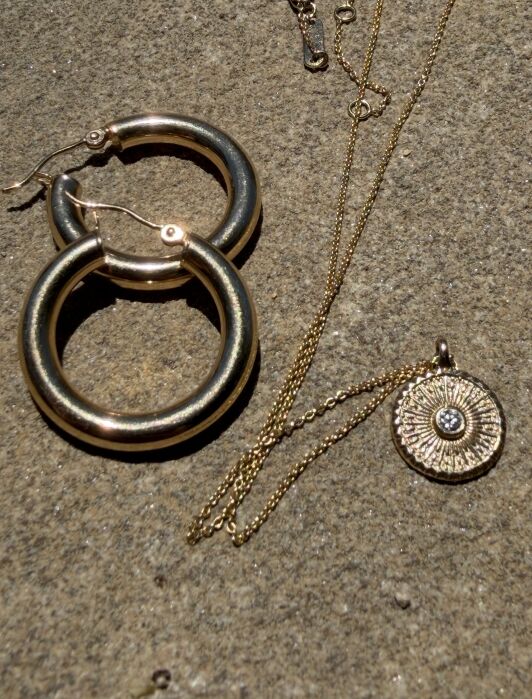 Two metallic hoop earrings and a gold necklace with a round, decorative pendant rest on a stone surface in sunlight.
