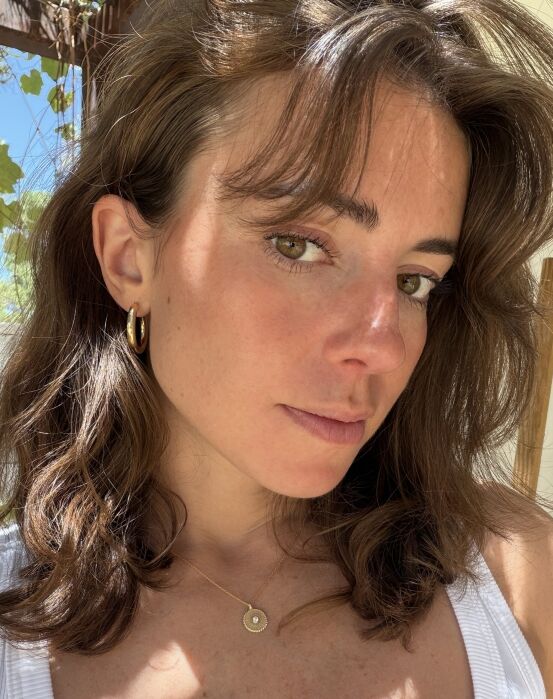 Woman with brown hair wearing a white top, gold hoop earring, and a pendant necklace, standing in sunlight with a leafy background.