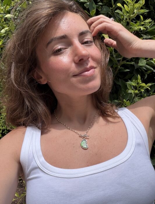 A person wearing a white tank top and a green pendant necklace takes a selfie outdoors, with greenery in the background and sunlight on their face.