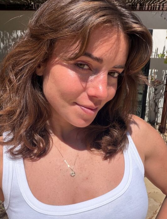A woman with wavy brown hair wearing a white tank top and a necklace stands outdoors in sunlight, looking at the camera.