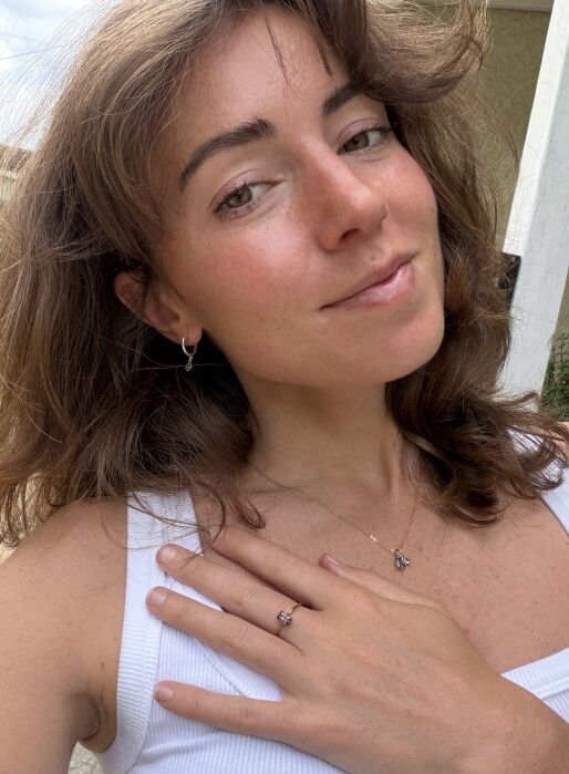 A woman with wavy brown hair wears a white tank top, a necklace, a ring, and hoop earrings, with one hand on her chest, smiling slightly at the camera.