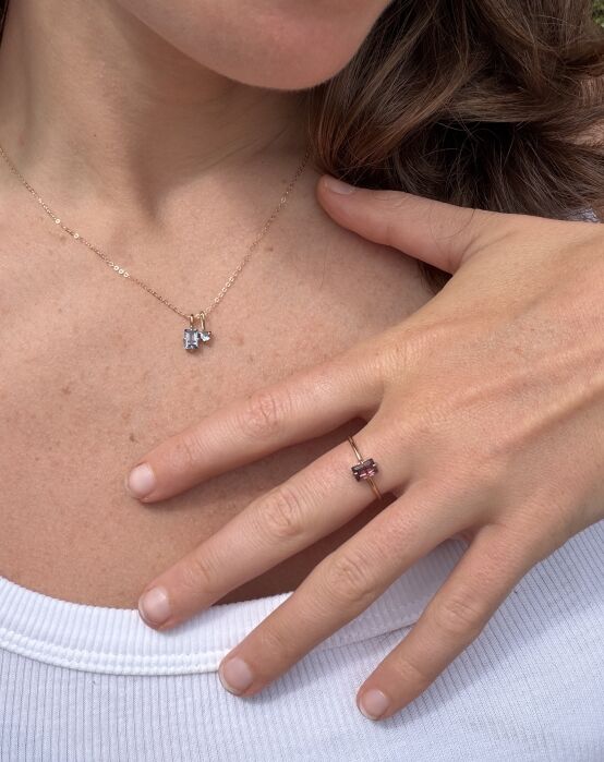 A person wearing a gold necklace with small charms and a gold ring with a rectangular pink stone, resting their hand on their chest over a white top.