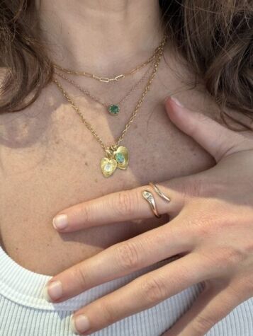 A person wearing layered gold necklaces with pendants and an emerald, along with a gold ring with a small stone, rests their hand on their chest over a white ribbed top.
