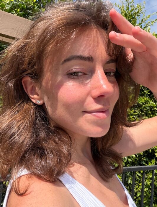 Woman with brown hair in a white tank top stands outside, tilting her head and holding one hand to her forehead in bright sunlight. Green foliage and a wooden structure are in the background.