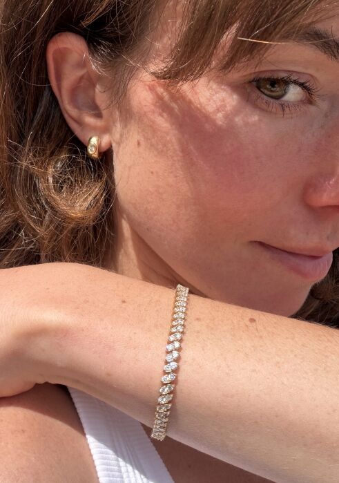 A woman wears a gold stud earring and a gold bracelet with diamonds, resting her arm on her face while looking slightly to the side.