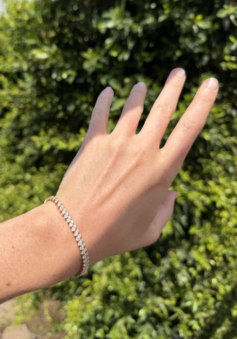 A hand wearing a thin diamond tennis bracelet is held out in front of a green leafy background in bright sunlight.