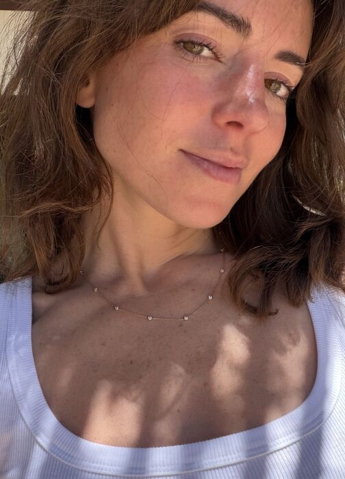 A person with wavy brown hair and green eyes wears a white tank top and a delicate necklace, standing in sunlight with shadows on their skin.