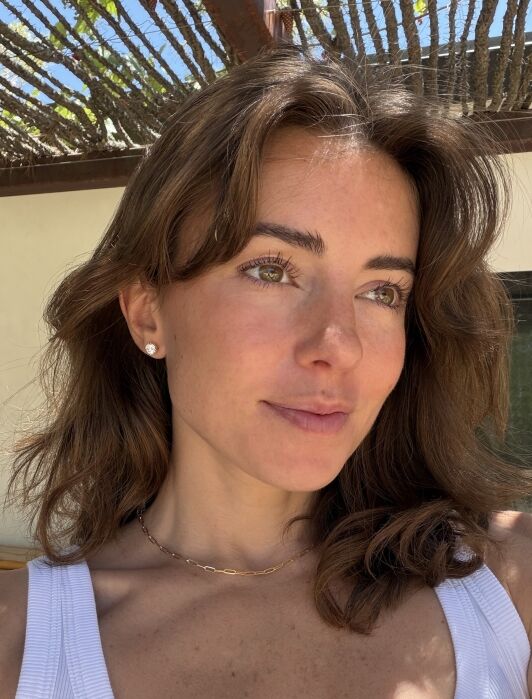 A woman with wavy brown hair and light eyes wears a white top and gold necklace, standing outside in natural sunlight with tree branches visible above her.