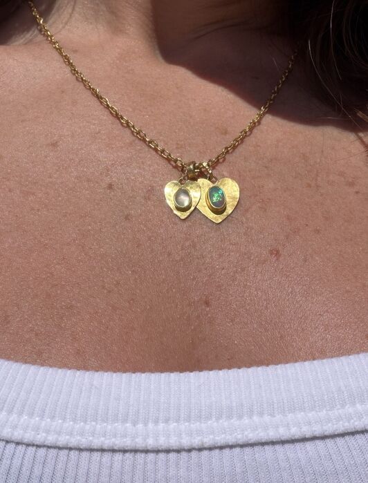 Gold chain necklace with two heart-shaped pendants, each featuring a different gemstone, worn on a person's neck above a white top.