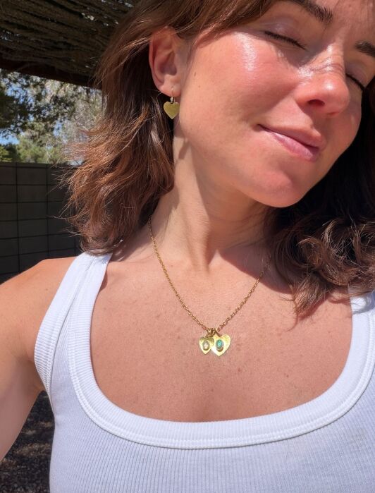 Woman in a white tank top outdoors, wearing gold heart-shaped earrings and a gold necklace with two avocado charms, eyes closed, smiling softly.
