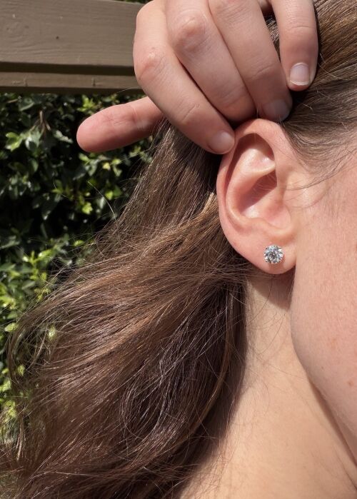 A person with long brown hair holds their hand near their ear, displaying a round, clear stud earring. Green foliage and a wooden structure are visible in the background.