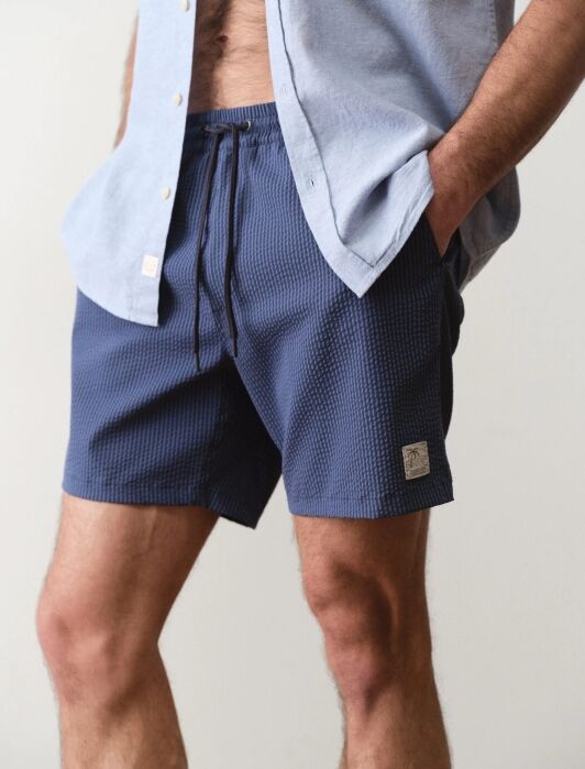 A person wearing a light blue button-up shirt and dark blue textured shorts with hands in pockets, standing against a plain background.