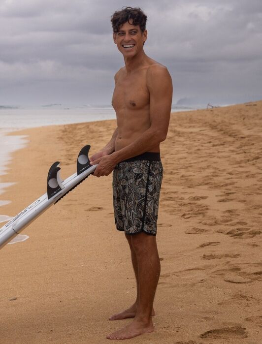 A man in patterned swim shorts stands barefoot on a sandy beach, holding a surfboard with cloudy skies overhead.