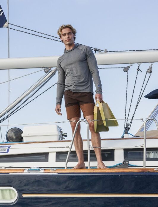 A man wearing a gray long-sleeve shirt and brown shorts stands barefoot on a boat, holding yellow swim fins, with rigging and equipment visible in the background.