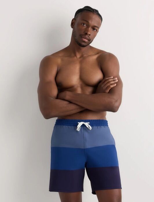 A man stands against a light gray wall, shirtless, wearing blue striped swim trunks with a white drawstring and his arms crossed.