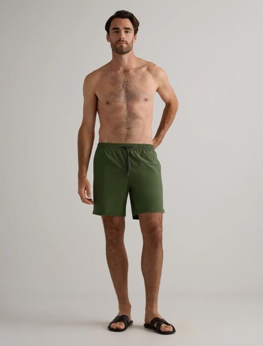 A shirtless man stands against a plain background wearing olive green shorts and black sandals.