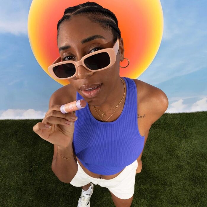 Woman wearing sunglasses, a blue tank top, and white shorts stands on grass, holding a lip balm with a bright orange sun and blue sky in the background.