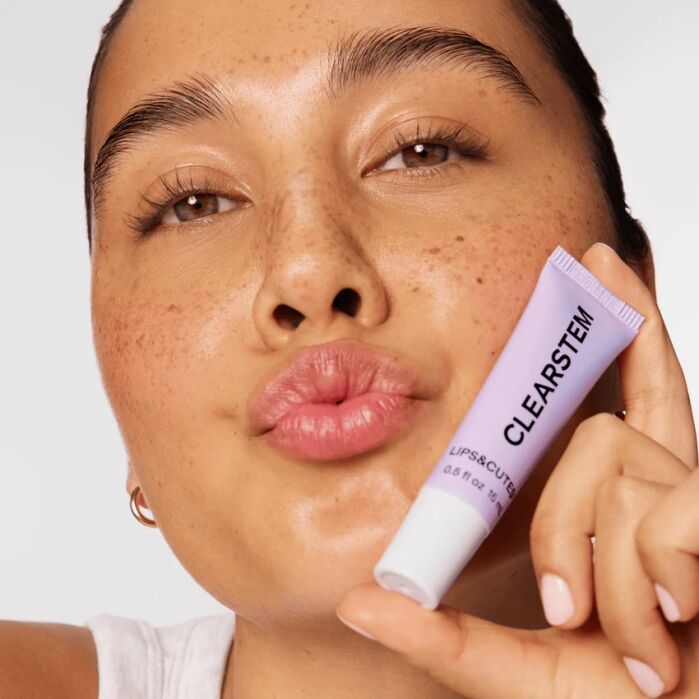 A person with freckles puckers their lips and holds a tube of CLEARSTEM LIPSTEM lip treatment near their face.