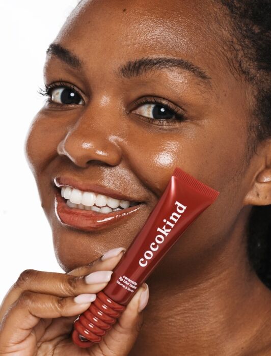 A smiling person with glowing skin holds a red tube labeled “cocokind” close to their face against a white background.