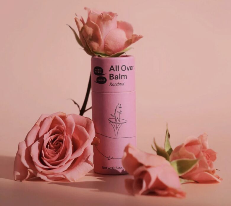 Pink tube labeled "All Over Balm Rosebud" stands upright, surrounded by three pink roses on a matching pink background.