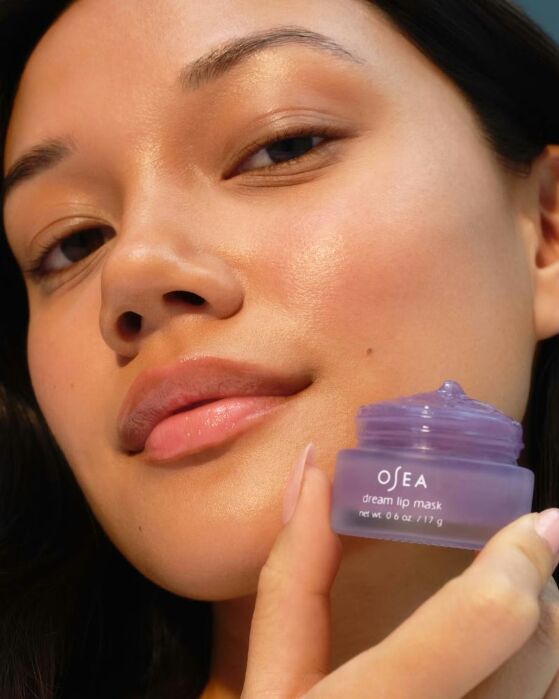 A person holds an open jar of OSEA Dream Lip Mask close to their face, showing smooth, glowing skin and natural lips.