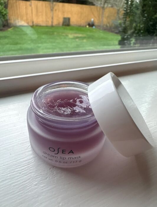 A jar of OSEA dream lip mask with the lid off sits on a white windowsill; a grassy yard and wooden fence are visible outside.