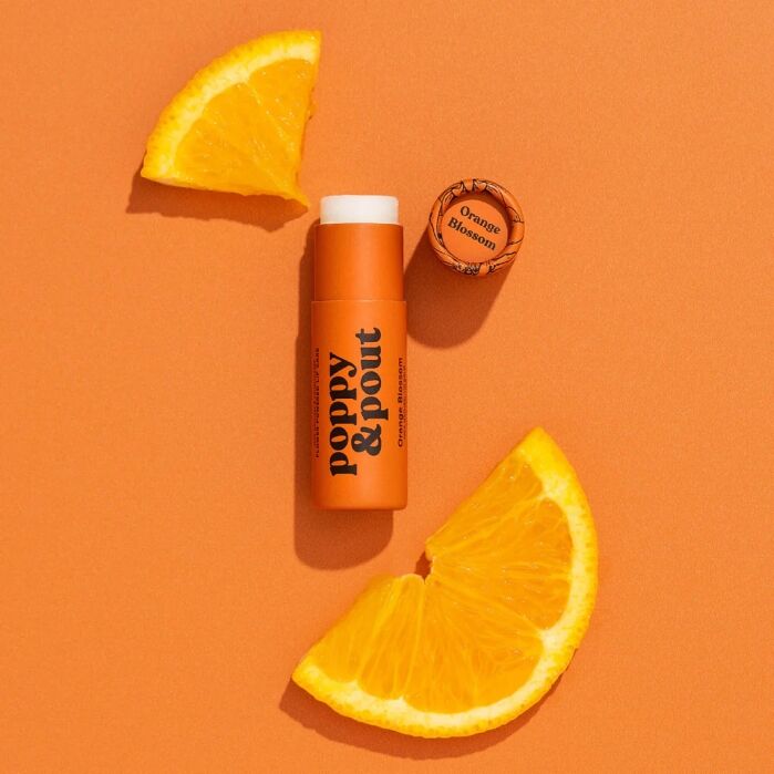 An orange tube of Poppy & Pout lip balm labeled "Orange Blossom" is surrounded by three orange slices on an orange background.