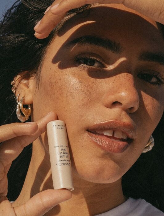 A person holds a tube of sun lip balm SPF 15 near their face, with sunlight and shadows falling across their skin.