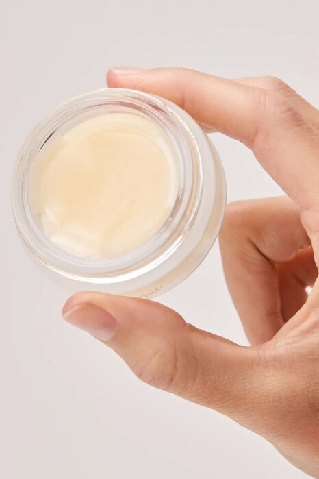A hand holding a small, clear jar filled with a light beige cream or balm, viewed from above against a plain background.