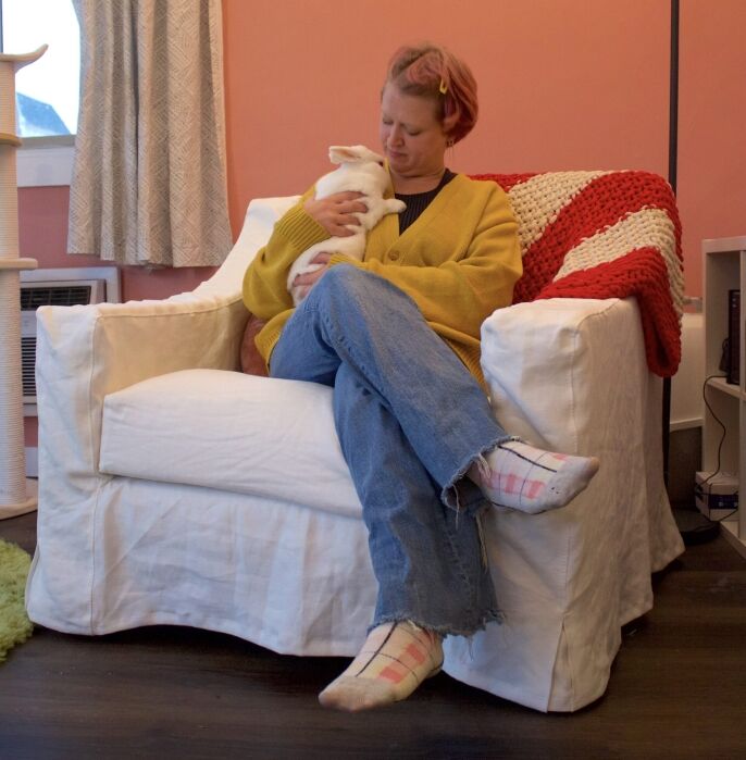 A person with short hair and a yellow cardigan sits on a white armchair, holding a white rabbit, in a cozy room with pink walls and a striped blanket.