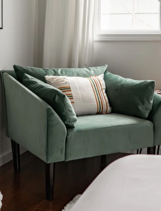 A green velvet armchair with two cushions and a striped pillow is placed in the corner of a room next to a small wooden side table and a window with white curtains.