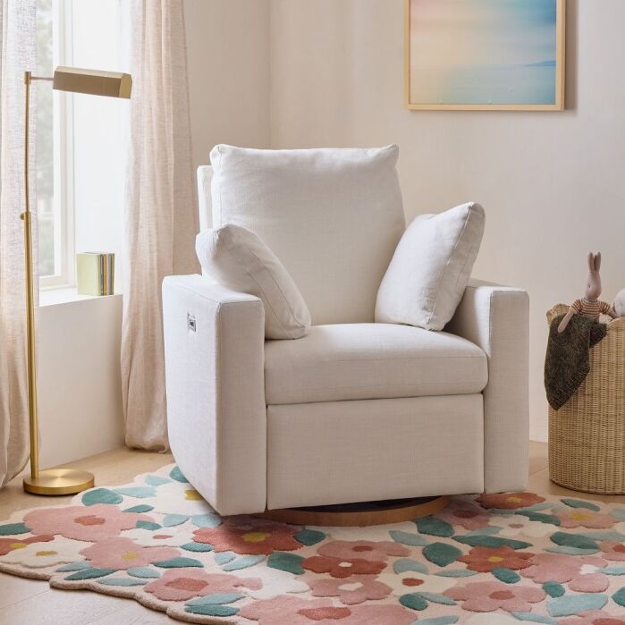A white armchair with two cushions sits on a colorful floral rug near a gold floor lamp and a woven basket in a softly lit room.