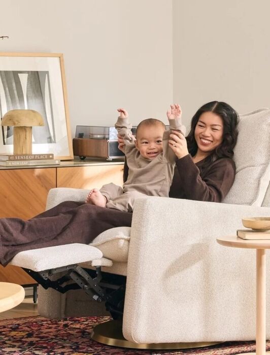 A woman and a young child sit together on a reclining armchair in a cozy, sunlit living room with modern decor and a patterned rug.
