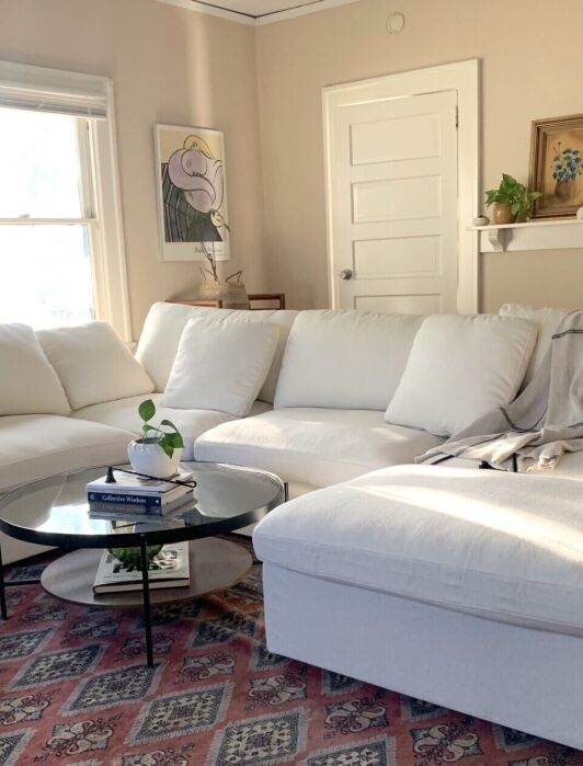 Bright living room with a large white sectional sofa, glass coffee table, patterned rug, and wall art near two doors. A gray blanket and small plant are on the table.