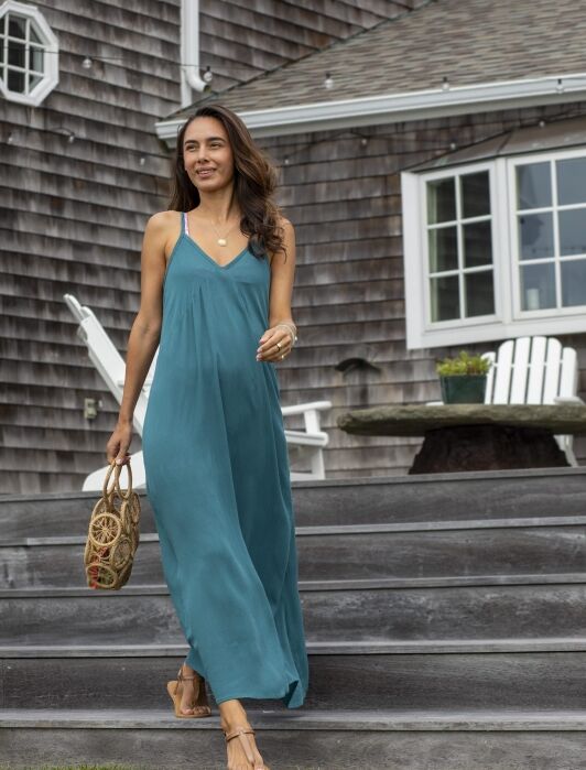 Woman in a teal maxi dress and sandals walks down wooden steps outside a shingle-style house, carrying a woven bag.