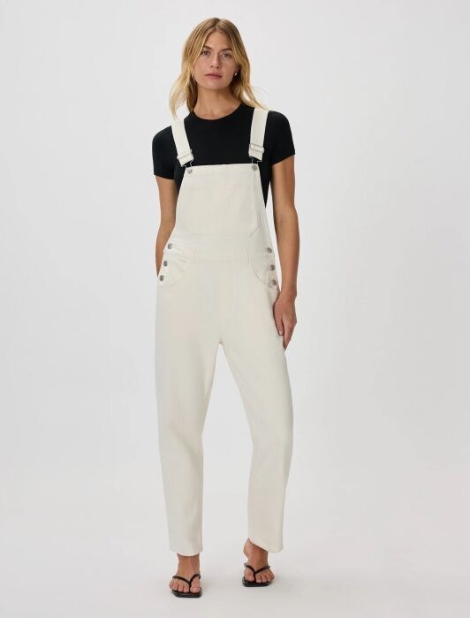 Person stands facing forward wearing a black t-shirt, white overalls, and black sandals against a plain white background.
