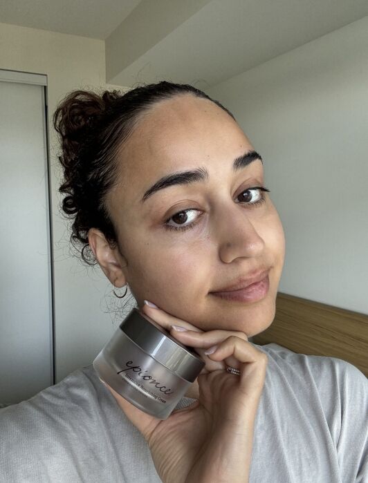 A person holds a jar of Epionce moisturizer next to their face, standing indoors against a plain background.