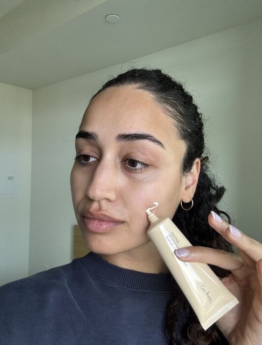 Person with dark hair holds a tube of cream, squeezing some onto their cheek in a well-lit room.