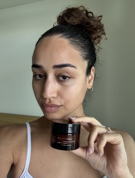 Woman with pulled-back hair and minimal makeup holds a jar of True Botanicals Chebula Extreme Cream, standing indoors with a neutral background.