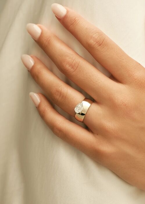 A hand with light skin and manicured nails wears a wide silver ring set with a teardrop-shaped clear gemstone, resting on a soft, white fabric background.