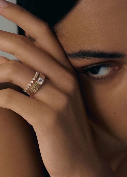 Close-up of a person’s face partially covered by their hand, wearing two gold rings, one with gemstones and the other with a textured design.