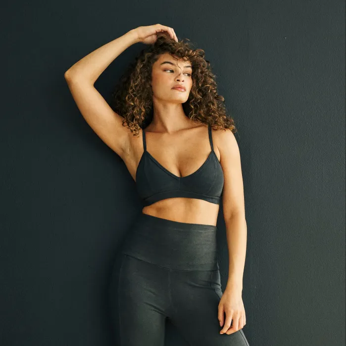 Woman with curly hair wearing a dark sports bra and high-waisted leggings poses against a dark wall with one arm raised above her head.