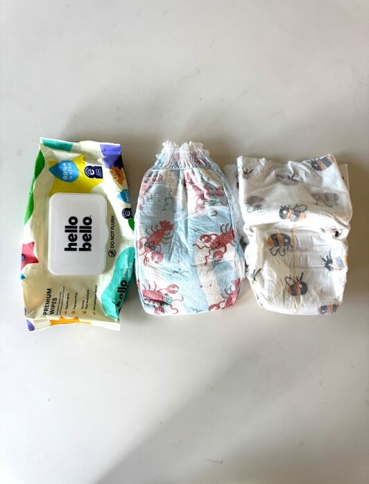 A pack of Hello Bello baby wipes, a pull-up style diaper with crab prints, and a folded disposable diaper are arranged side by side on a white surface.
