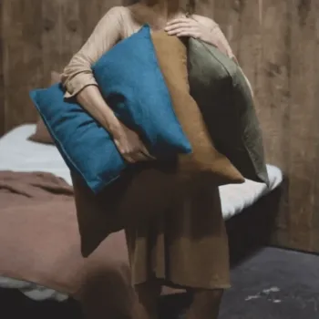 A person in a brown dress stands in a bedroom holding three large pillows—one blue, one brown, and one green—against a background of a bed and wooden wall.
