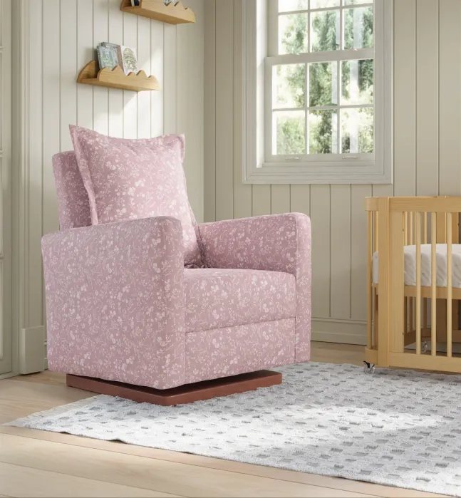 A pink floral armchair with matching pillow sits on a patterned rug beside a wooden crib in a sunlit nursery with beige paneled walls and wall shelves.