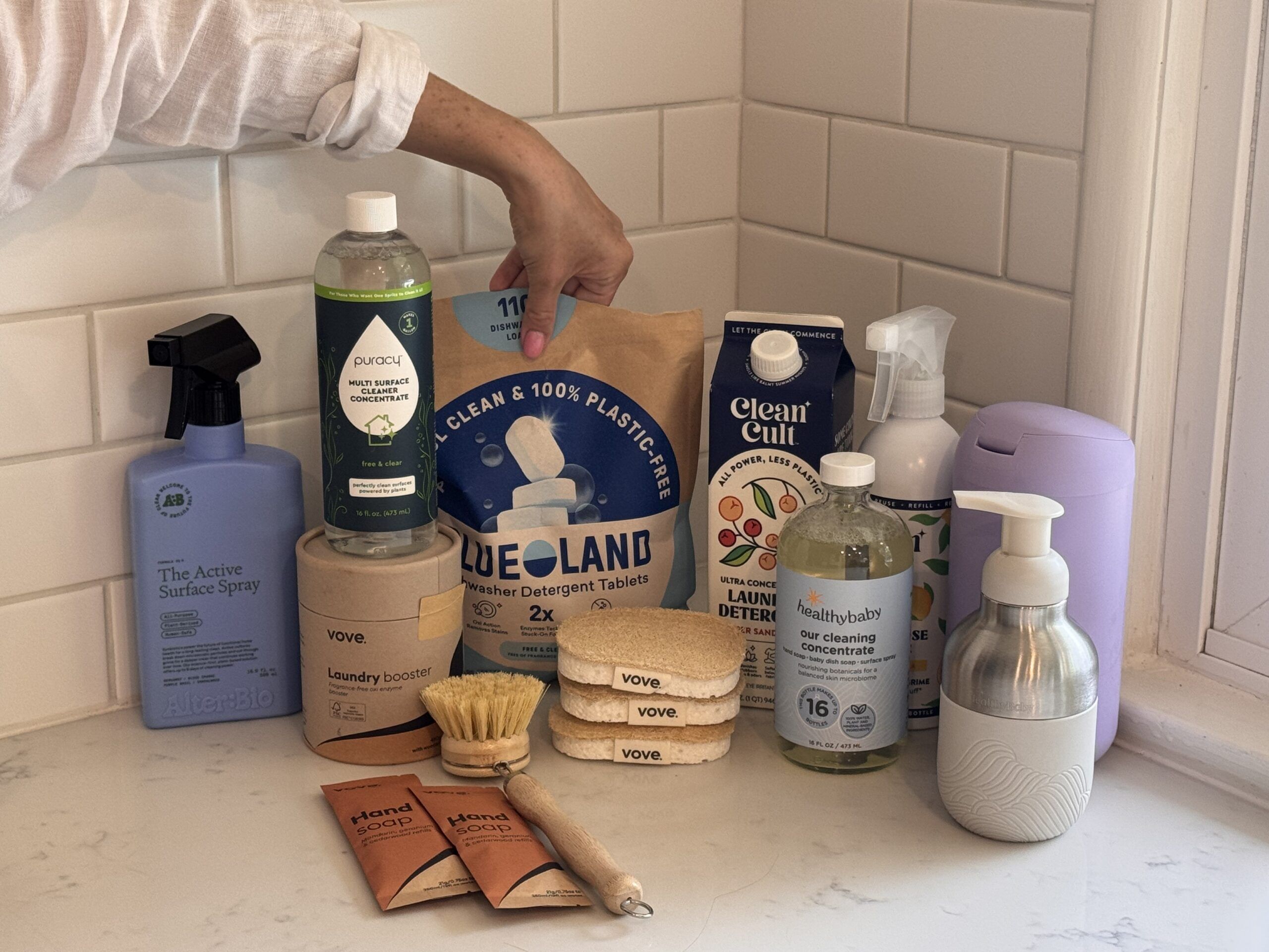 A person arranges eco-friendly cleaning and laundry products, including detergent, soap, wipes, sprays, and brushes, on a white bathroom counter near a sink.